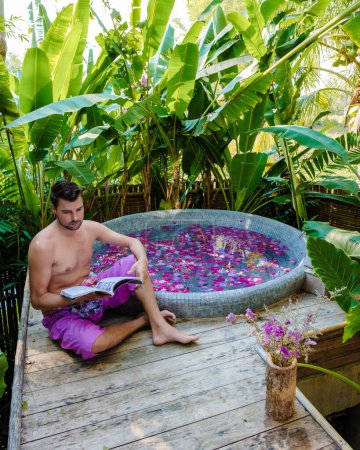 Téléchargez les photos : Hommes lisant un livre dans une baignoire dans la forêt tropicale de Thaïlande pendant des vacances avec des fleurs dans le bain - en image libre de droit