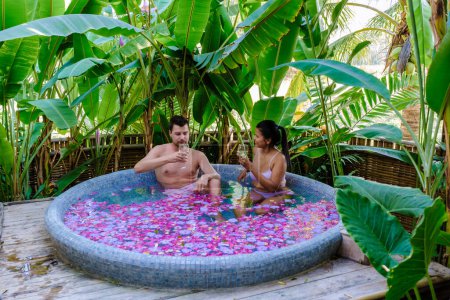 Foto de Pareja en una bañera en la selva tropical de Tailandia durante unas vacaciones con flores en el baño, hombres y mujeres relajarse en un jacuzzi con pétalos de rosa en el exterior en la naturaleza. Celebración de San Valentín - Imagen libre de derechos