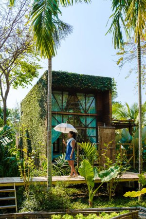 Photo for Asian women walk with umbrella at a wooden cottage surrounded by palm trees and a vegetable garden in the countryside. cabin in tropical rainforest - Royalty Free Image