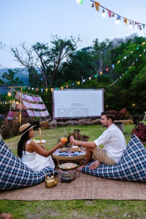 Téléchargez les photos : Couple hommes et femmes regardant un film dans le jardin d'un film de cinéma en plein air dans un jardin tropical avec des lumières de Noël. H - en image libre de droit