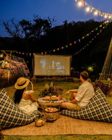 Ein Paar Männer und Frauen schaut sich im Garten eines Outdoor-Kinos in einem tropischen Garten mit Weihnachtsbeleuchtung einen Film an. H