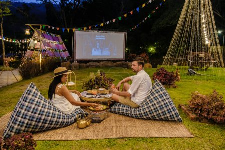 Téléchargez les photos : Couple hommes caucasiens et femmes asiatiques regardant un film dans le jardin d'un film de cinéma en plein air dans un jardin tropical avec des lumières de Noël. H - en image libre de droit
