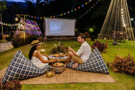 Téléchargez les photos : Couple hommes et femmes regardant un film dans le jardin d'un film de cinéma en plein air dans un jardin tropical avec des lumières de Noël. H - en image libre de droit