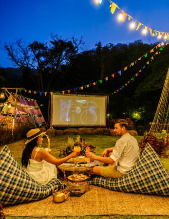 Téléchargez les photos : Couple hommes caucasiens et femmes asiatiques regardant un film dans le jardin d'un film de cinéma en plein air dans un jardin tropical avec des lumières de Noël. H - en image libre de droit