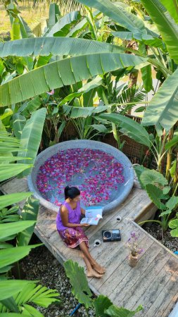 Foto de Mujeres relajándose en una bañera en la selva tropical de Tailandia durante unas vacaciones con flores en el baño, mujeres relajándose en un jacuzzi con pétalos de rosa afuera en la naturaleza leyendo un libro - Imagen libre de derechos