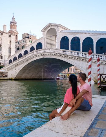 Téléchargez les photos : Un couple d'hommes et de femmes en voyage à Venise en Italie assis au bord de l'eau du pont du Rialto à Venise, Italie. Architecture et point de repère de Venice.cityscape de Venise Italie pendant l'été - en image libre de droit