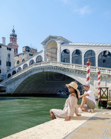 Téléchargez les photos : Un couple d'hommes et de femmes en voyage à Venise Italie assis au bord de l'eau du pont du Rialto à Venise, Italie. - en image libre de droit