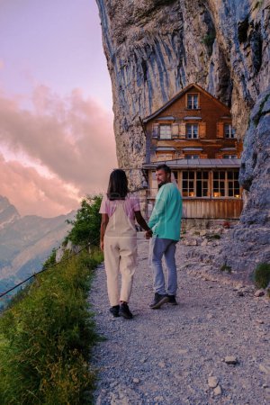 Téléchargez les photos : Couple d'hommes et de femmes randonnée à Berggasthaus Aescher en den Appenzeller Alpen au coucher du soleil, restaurant sous une falaise sur la montagne Ebenalp en Suisse, Appenzell - en image libre de droit