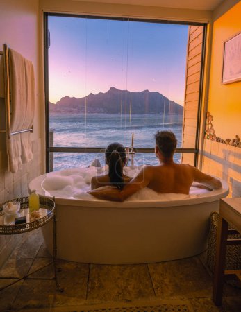 Foto de Couple man and women in the bathtub looking out over the ocean of Cape Town South Africa during vacation. Bath Tub during sunset, man and woman in bath tub jaccuzi on vacation - Imagen libre de derechos