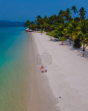 Téléchargez les photos : Un couple d'hommes et de femmes en vacances à Koh Mook île tropicale dans la mer d'Andaman en Thaïlande, hommes et femmes marchant sur une plage avec des palmiers - en image libre de droit
