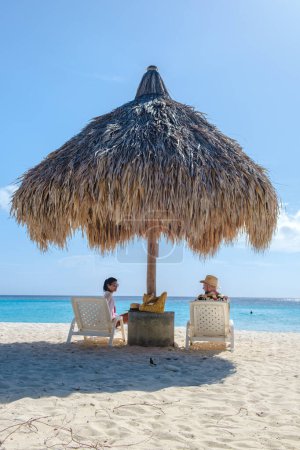 Foto de Un par de hombres y mujeres de vacaciones en Curazao, Cas Abao Beach Playa Cas Abao Caribe isla de Curazao, Playa Cas Abao tropical playa blanca con sillas de playa y un paraguas de playa tatuado - Imagen libre de derechos