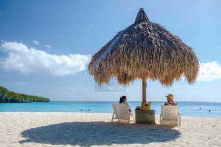 A couple of men and women on vacation in Curacao, Cas Abao Beach Playa Cas Abao Caribbean island of Curacao, Playa Cas Abao tropical white beach with beach chairs and a tatched beach umbrella 