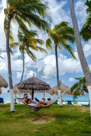 Photo for Palm Beach Aruba Caribbean, a couple of men and women in a hammock at a white long sandy beach with palm trees at Aruba Antilles. - Royalty Free Image