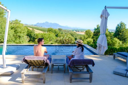 Téléchargez les photos : Couple d'hommes et de femmes se relaxant dans une piscine avec vue sur un paysage viticole au coucher du soleil avec des montagnes à Stellenbosch, près de Cape Town, Afrique du Sud. - en image libre de droit