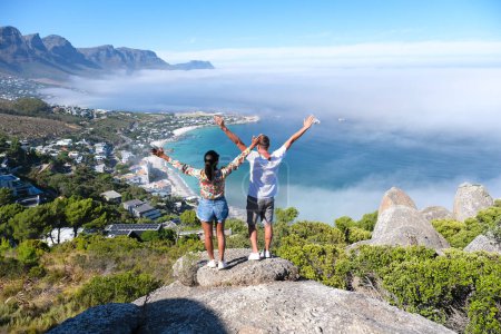 Foto de Un par de hombres y mujeres con las manos en alto saliendo en el mirador de The Rock en Ciudad del Cabo sobre Campsbay, vista sobre Camps Bay con niebla sobre el océano. niebla procedente del océano Ciudad del Cabo Sudáfrica - Imagen libre de derechos