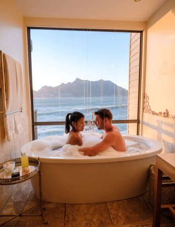 Téléchargez les photos : Un couple d'hommes et de femmes dans une baignoire donnant sur l'océan du Cap en Afrique du Sud pendant les vacances. Baignoire au coucher du soleil, jacuzzi en vacances de luxe - en image libre de droit