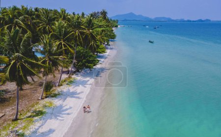 Téléchargez les photos : Un couple d'hommes et de femmes en vacances à Koh Mook île tropicale dans la mer d'Andaman en Thaïlande, hommes et femmes marchant sur une plage avec des palmiers drone vue aérienne - en image libre de droit