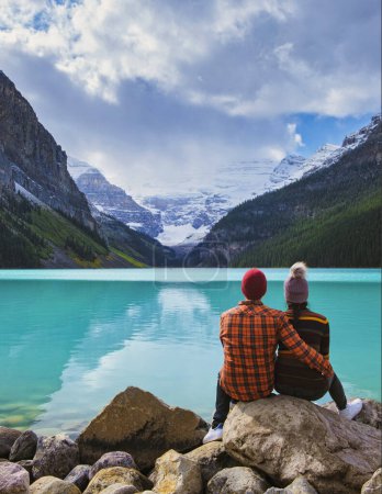 Foto de Lake Louise Banff national park is a lake in the Canadian Rocky Mountains. A young couple of men and women sitting on a rock by the lake during a cold day in Autumn in Canada - Imagen libre de derechos