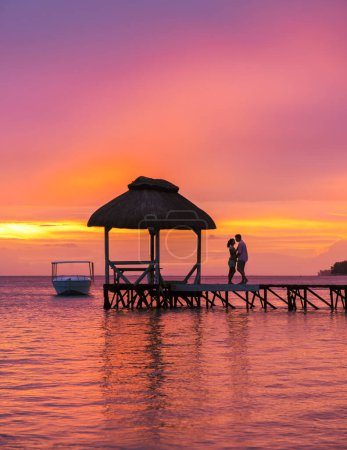 Téléchargez les photos : Homme et femme regardant le coucher du soleil sur une jetée en bois dans l'océan sur une plage tropicale à Maurice, un couple en vacances de lune de miel à Maurice - en image libre de droit