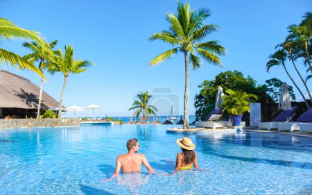 Téléchargez les photos : Homme et Femme se relaxant dans une piscine, un couple en vacances de lune de miel à Maurice bronzant dans la piscine avec palmiers et chaises longues - en image libre de droit