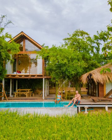 Photo for A couple of men and women in front of a Bamboo hut homestay farm, with Green rice paddy fields in Central Thailand with a small plunge pool - Royalty Free Image
