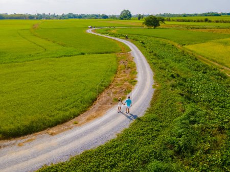 Foto de Un par de hombres y mujeres de vacaciones en Tailandia caminando por un sinuoso camino rural curvado en medio de arrozales de arroz verde en la región central de Tailandia Suphanburi, vista aérea de drones - Imagen libre de derechos