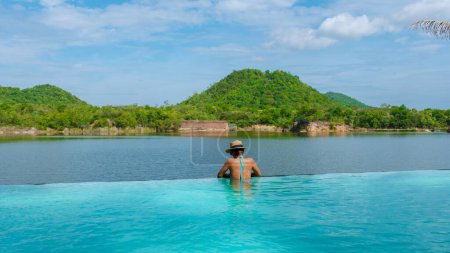 Téléchargez les photos : Femmes asiatiques au bord d'une piscine en vacances en Thaïlande donnant sur le lac et les montagnes verdoyantes de la Thaïlande centrale. - en image libre de droit