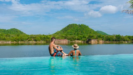 Téléchargez les photos : Couple sur le bord d'une piscine en vacances en Thaïlande donnant sur le lac et les montagnes verdoyantes de la Thaïlande centrale. - en image libre de droit