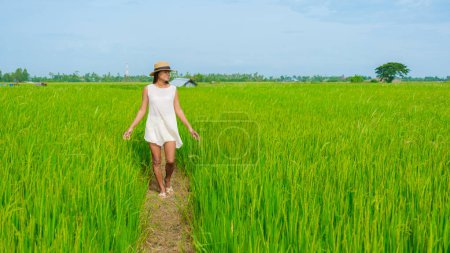 Photo for Aian woman on vacation in Thailand walking on a countryside road in the middle of green rice paddy fields in Central Thailand Suphanburi region, drone aerial view of green rice fields in Thailand - Royalty Free Image