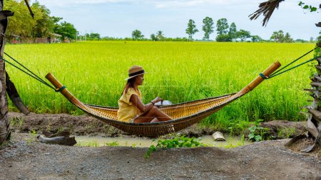 Téléchargez les photos : Femmes asiatiques dans un hamac travaillant sur un ordinateur portable avec sur le fond des rizières vertes en Thaïlande - en image libre de droit