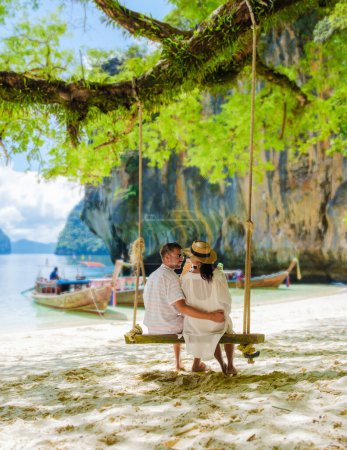 Téléchargez les photos : Koh Lao Lading près de Koh Hong Krabi Thaïlande, est une belle plage avec des bateaux à queue longue, un couple d'hommes européens, et une femme asiatique sur la plage se détendre sur une balançoire sous un arbre. Couple en excursion en bateau - en image libre de droit