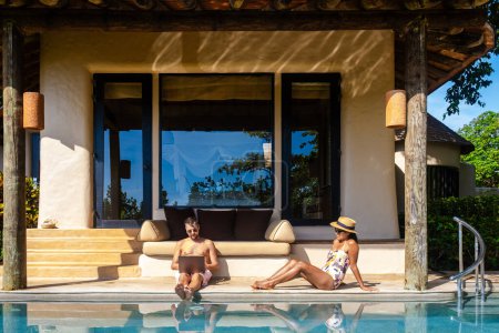 Téléchargez les photos : Hommes travaillant sur un ordinateur portable un nomade numérique travaillant dans une piscine pendant des vacances sur une île tropicale. homme et femme dans la piscine à débordement au coucher du soleil. vacances de luxe dans une villa piscine de luxe - en image libre de droit