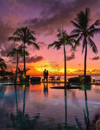 Foto de Un par de hombres y mujeres mirando el atardecer en una playa tropical en Mauricio con palmeras junto a la piscina, atardecer tropical en la playa en Mauricio. - Imagen libre de derechos