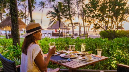 Téléchargez les photos : Dîner romantique sur la plage à Koh Kood Thaïlande, femme regardant coucher de soleil pendant le dîner sur la plage avec une jetée en bois et patio privé sur des vacances de luxe en Thaïlande - en image libre de droit