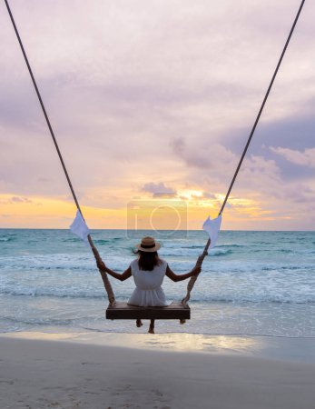Foto de Mujer tailandesa asiática en un columpio de cuerda bajo una palmera observando la puesta de sol en la playa de Koh Kood Island Tailandia Trat, playa tropical con palmeras y un océano de color turquesa - Imagen libre de derechos