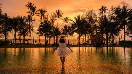 Téléchargez les photos : Femme asiatique regardant le coucher du soleil dans une piscine tropicale avec des palmiers sur l'île de Koh Kood Thaïlande, piscine pendant le coucher du soleil sur la plage dans un hôtel de luxe - en image libre de droit