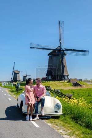 Photo for Couple doing a road trip with an old vintage car in the Dutch flower windmill region, men and woman at an old retro vintage car with on the background historical wooden windmills - Royalty Free Image