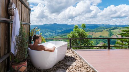 Téléchargez les photos : Un homme dans une baignoire donnant sur les montagnes de Chiang Rai nord de la Thaïlande pendant les vacances. Salle de bain extérieure, jeunes hommes dans une baignoire pendant le coucher du soleil - en image libre de droit
