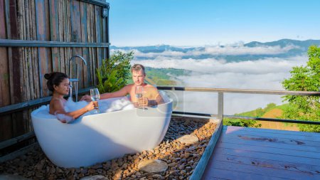 Téléchargez les photos : Un couple d'hommes et de femmes dans une baignoire donnant sur les montagnes de Chiang Rai nord de la Thaïlande pendant les vacances. Salle de bain extérieure, baignoire au lever du soleil - en image libre de droit