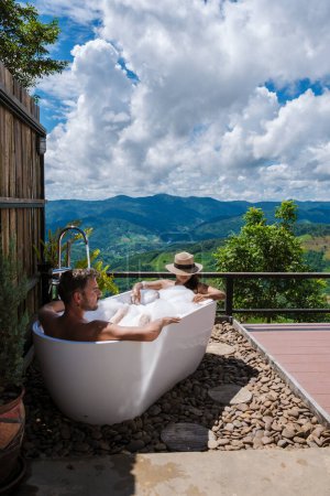 Téléchargez les photos : Un couple d'hommes et de femmes dans une baignoire donnant sur les montagnes de Chiang Rai nord de la Thaïlande pendant les vacances. Salle de bain extérieure, baignoire au lever du soleil - en image libre de droit