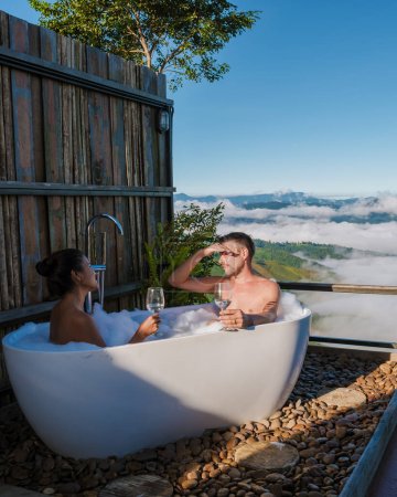 Photo for A multiracial mixed couple of men and women in a bathtub looking out over the mountains of Chiang Rai Northern Thailand during vacation. Outdoor bathroom, bathtub during sunset - Royalty Free Image
