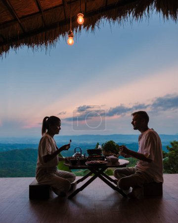 Téléchargez les photos : Un couple d'hommes et de femmes en vacances dans le nord de la Thaïlande, séjournant dans une cabane chez l'habitant dans les montagnes de Chiang Rai Doi Chang profiter d'un dîner avec un barbecue thaïlandais - en image libre de droit