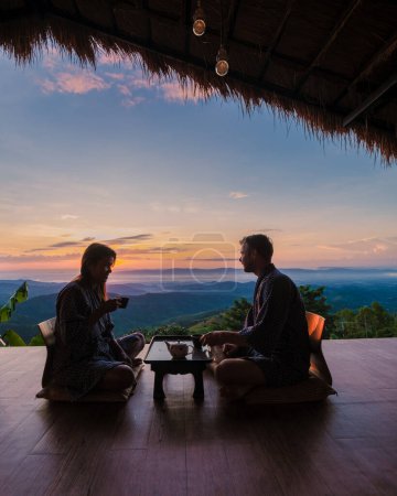 Photo for A couple of men and women drinking tea on the wooden balcony at sunrise, on vacation in Northern Thailand staying at a homestay cabin hut in the mountains of Chiang Rai Doi Chang - Royalty Free Image
