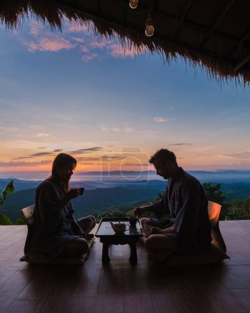 Téléchargez les photos : Un couple d'hommes et de femmes buvant du thé au lever du soleil en vacances dans le nord de la Thaïlande séjournant dans une cabane chez l'habitant dans les montagnes de Chiang Rai Doi Chang boire du thé sur le balcon en bois au lever du soleil - en image libre de droit