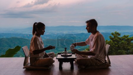 Photo for A couple of men and women drinking tea at sunrise on vacation in Northern Thailand staying at a homestay cabin hut in mountains of Chiang Rai Doi Chang drinking tea on the wooden balcony at sunrise - Royalty Free Image