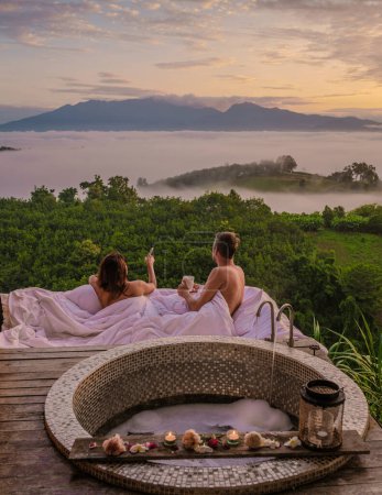 Téléchargez les photos : Un couple d'hommes et de femmes dans un lit extérieur donnant sur les montagnes du nord de la Thaïlande pendant les vacances. Salle de bain extérieure et baignoire au coucher du soleil dans les montagnes avec brouillard et brouillard - en image libre de droit