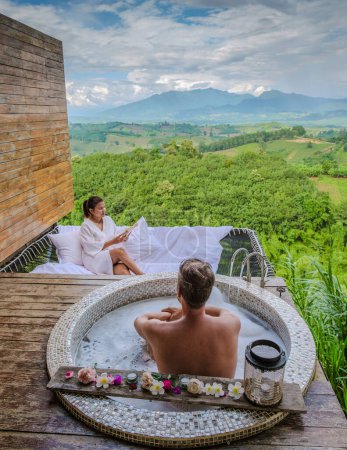 Téléchargez les photos : Un couple d'hommes et de femmes dans une baignoire donnant sur les montagnes du nord de la Thaïlande pendant les vacances. Salle de bain extérieure et baignoire au coucher du soleil dans les montagnes - en image libre de droit