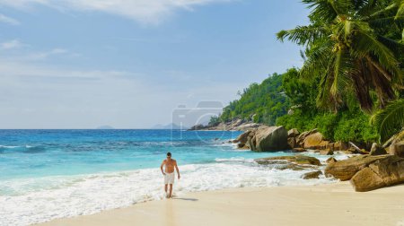 Téléchargez les photos : Petite Anse Beach Seychelles, un jeune homme en short de bain sur une plage tropicale blanche avec des palmiers Petite Anse beach Mahe Tropical Seychelles Islands. homme marchant sur la plage pendant les vacances - en image libre de droit