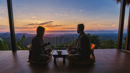 Téléchargez les photos : Un couple d'hommes et de femmes en vacances dans le nord de la Thaïlande, séjournant dans une cabane chez l'habitant dans les montagnes de Chiang Rai Doi Chang boire du thé sur le balcon en bois au lever du soleil - en image libre de droit