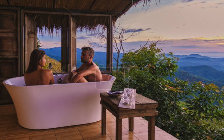 Photo for A mixed multiracial couple of men and women in a bathtub looking out over the mountains of Chiang Rai Northern Thailand during vacation. Outdoor bathroom, bathtub during sunset - Royalty Free Image